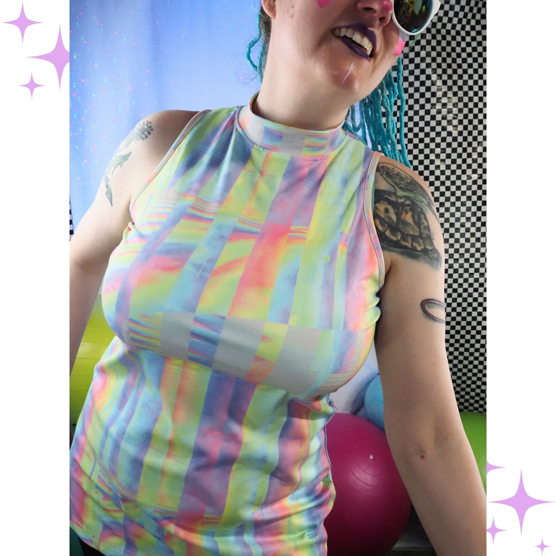 A woman wearing a sleeveless mock-neck top with a pastel rainbow holographic stripe pattern, set against a gym background with a pink exercise ball.