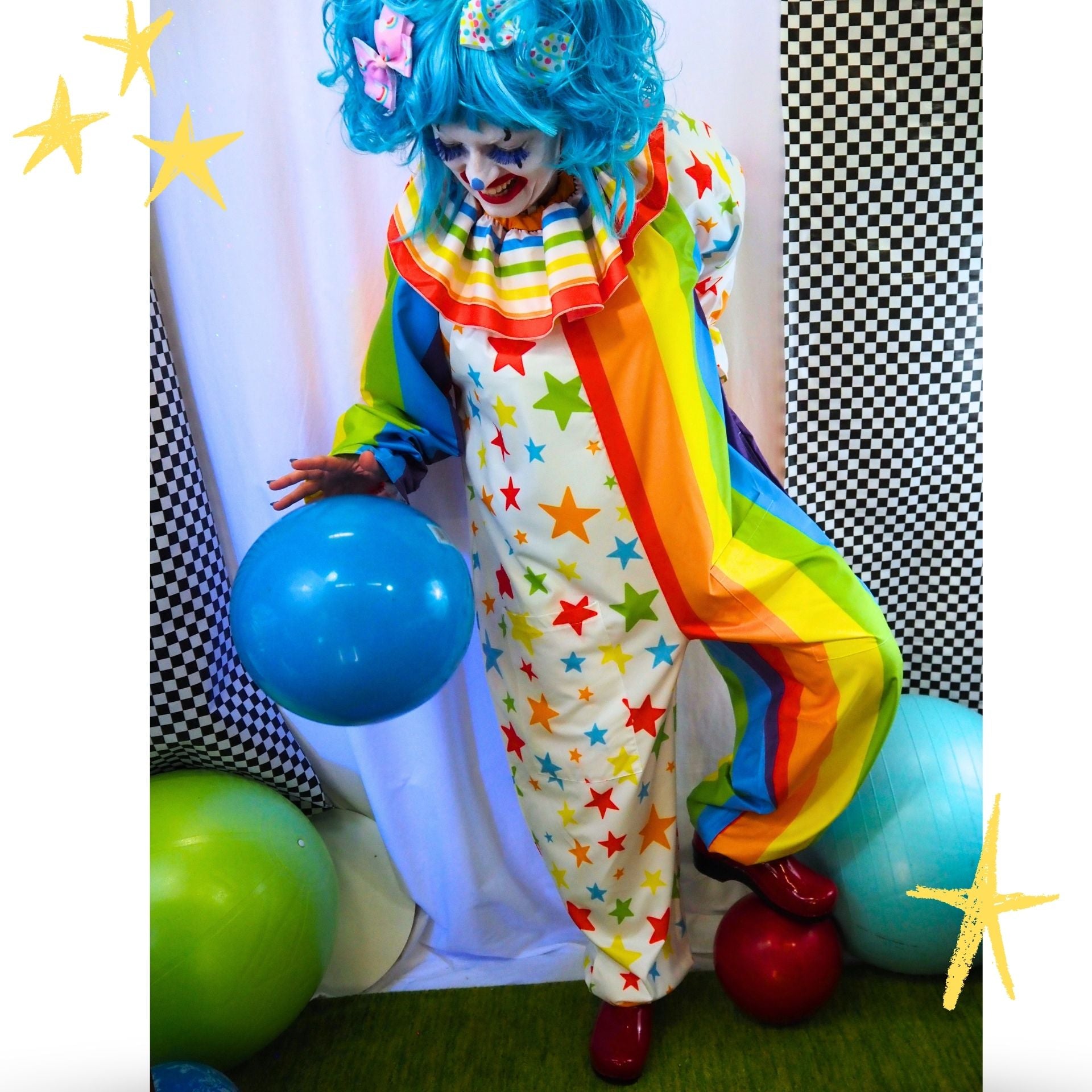 A person dressed as a clown wearing a white one-piece costume covered in colorful star patterns and rainbow-striped ruffles, with bright blue hair, holding a blue balloon in a decorated studio setting.