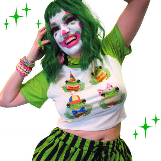 Person with clown makeup and green hair wearing a white crop top with colorful frog designs and green and black striped pants.