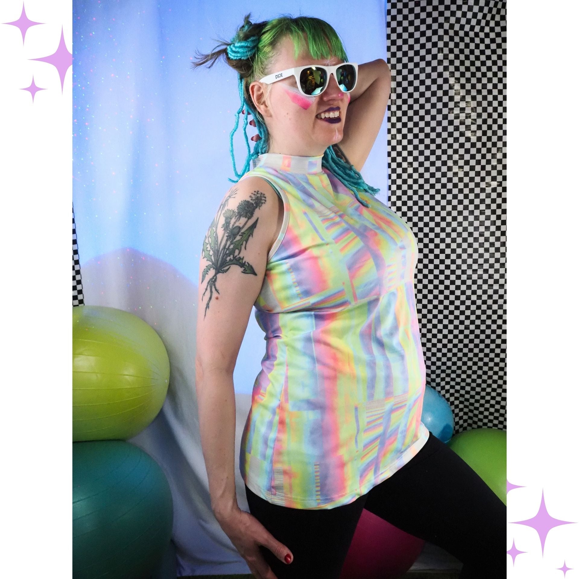A woman modeling a sleeveless, high-neck, form-fitting top with a pastel rainbow glitch pattern. She is wearing black leggings, white sunglasses, and has green hair in a bun, posing in front of a blue and black-and-white checkered backdrop with exercise balls.