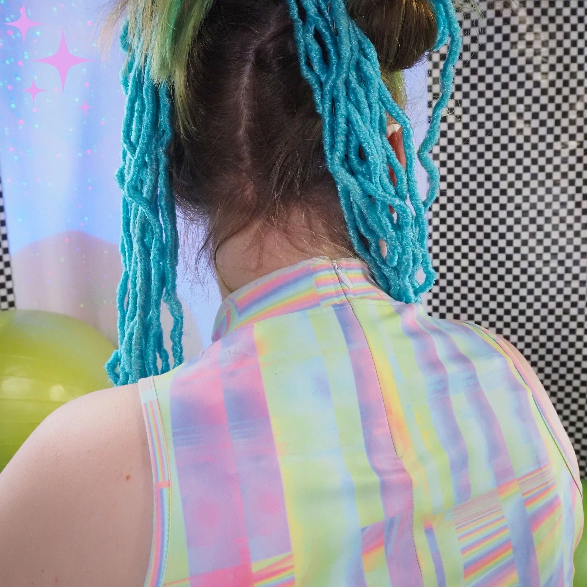 Back view of a person with bright turquoise dreadlocks and a pastel rainbow striped sleeveless top, standing indoors against a checkered wall and starry backdrop.