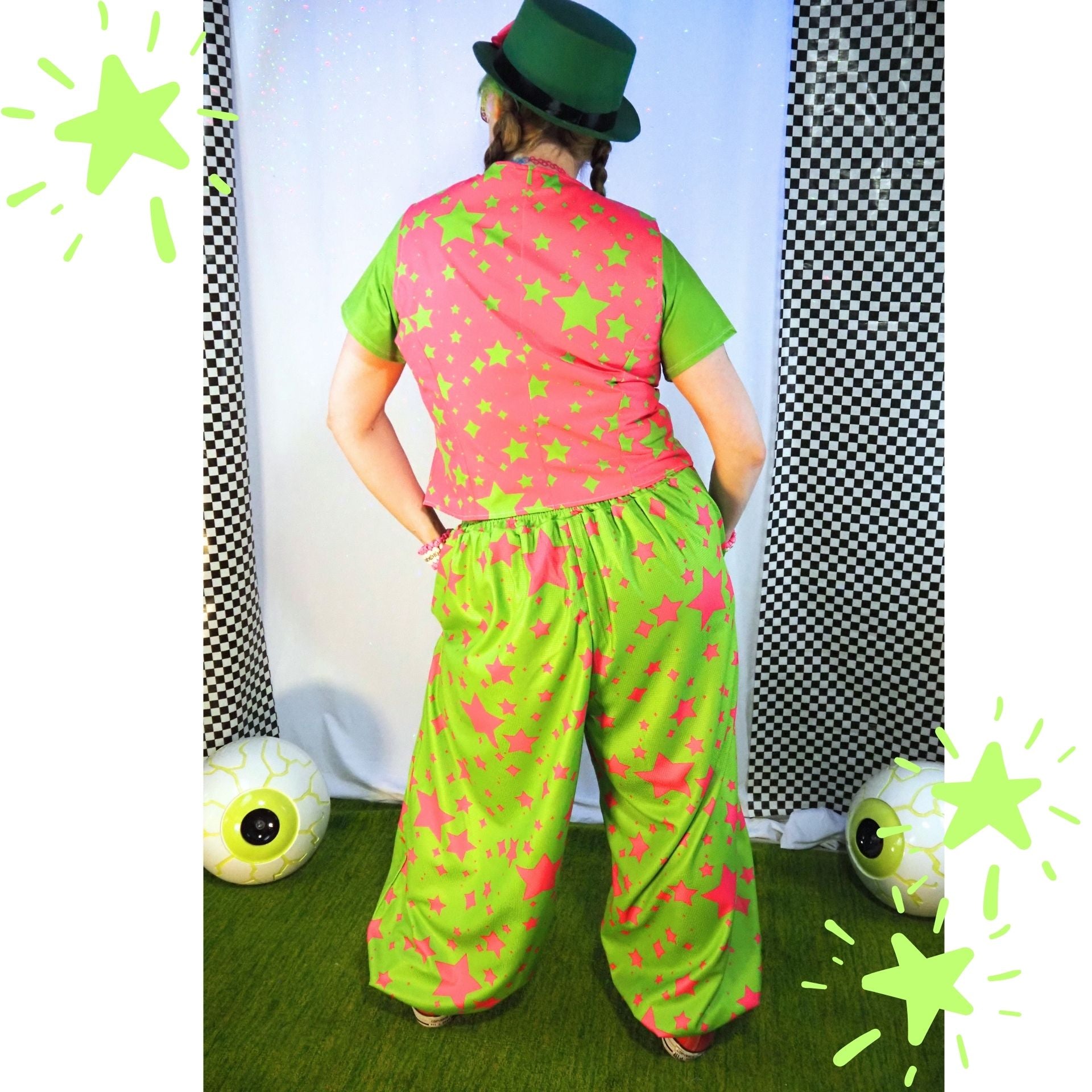 Person wearing a colorful outfit with a pink shirt and green pants, standing against a checkered curtain background.
